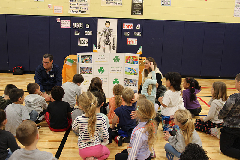 A large trifold board with many items about Ireland. A woman and man sit near it and a large group of young elementary age students listen and learn about Ireland.