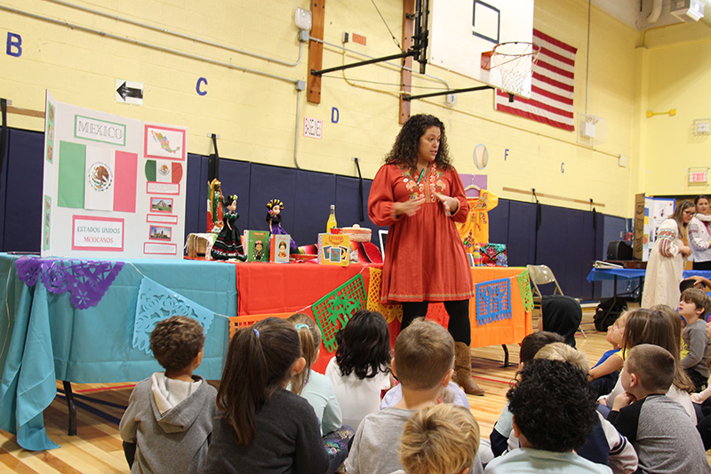 A woman in a red dress stands in front of a group of young elementary students talking about her native land of Mexico.