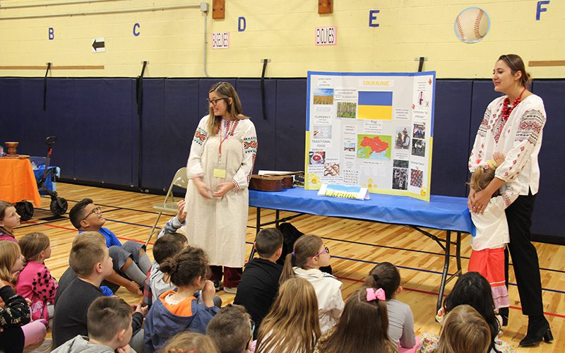 Two women dressed in traditional Ukrainian clothing, talk to a group of elementary students about Ukraine. There is a table with a large poster on it with facts about Ukraine.