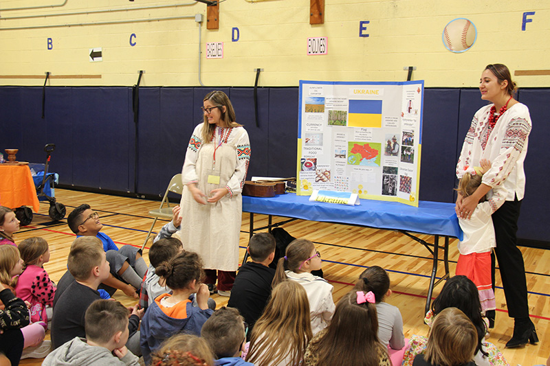 Two women dressed in traditional Ukrainian clothing, talk to a group of elementary students about Ukraine. There is a table with a large poster on it with facts about Ukraine.