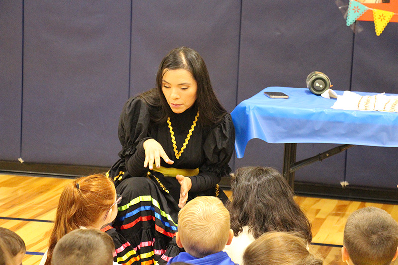 A woman with long dark hair, wearing a multi-colored striped skirt and black shirt, talks to a group of elementary students about Mexico.