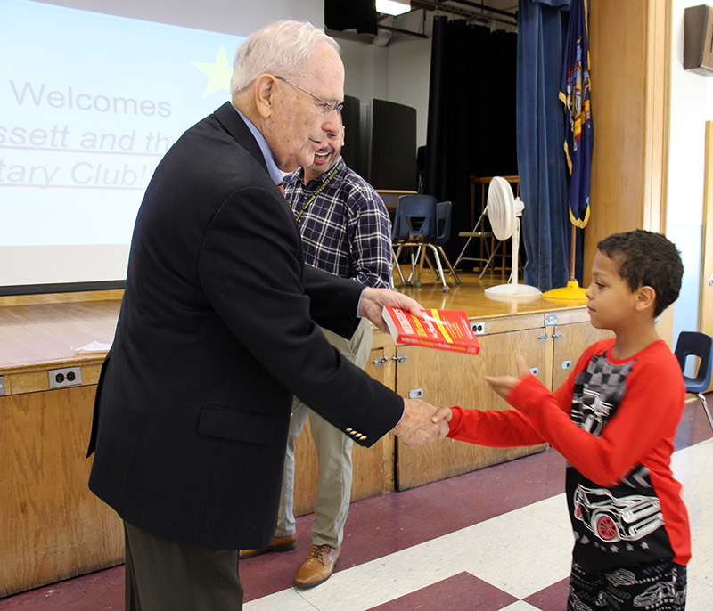 A man in a black suit jacket hands a red dictionary to a third-grade boy.
