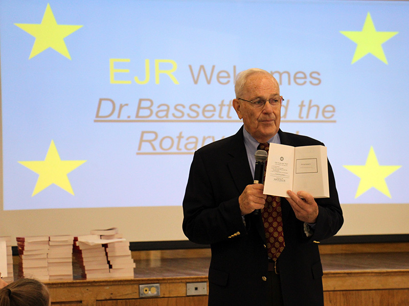 A man in a black suit jacket and tie holds up an open dictionary 