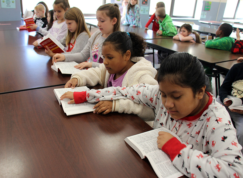 A line of third-graders sitting at a table look in the dictionaries they just received.