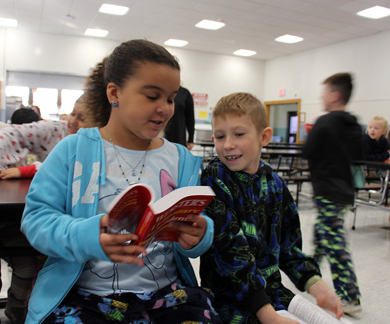 A third grade girl holds open a red dictionary while a boy sitting next to her reads it too.