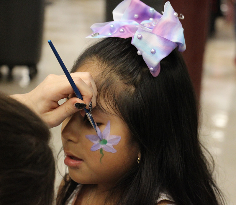 A girl with long dark hair and a multi-colored blow in her hair closes her eyes as someone paints a flower on her cheek.