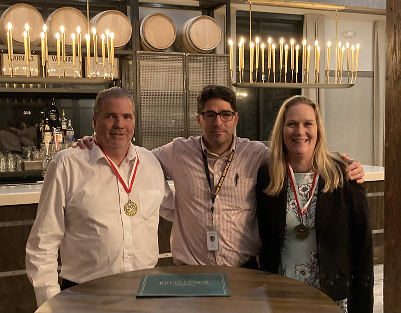 Three adults, two men and a woman on the right, smile. The man in the center, with dark hair and glasses, has an arm around each of the two, who are wearing medals around their necks.