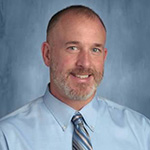 A man in a light blue shirt and blue striped tie smiles. His hair is very close cut.