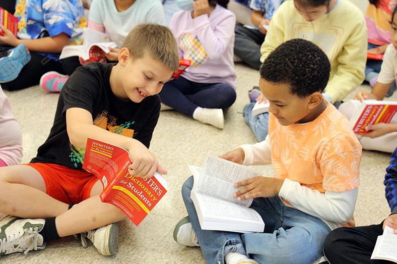 Two third-grade boys sit on the floor with their dictionaries open and smile as they look in them.