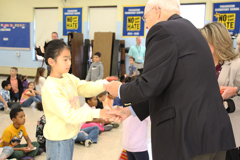 A third-grade girl in a yellow shirt shakes hands with a man in a black suit jacket and accepts a red dictionary from him.