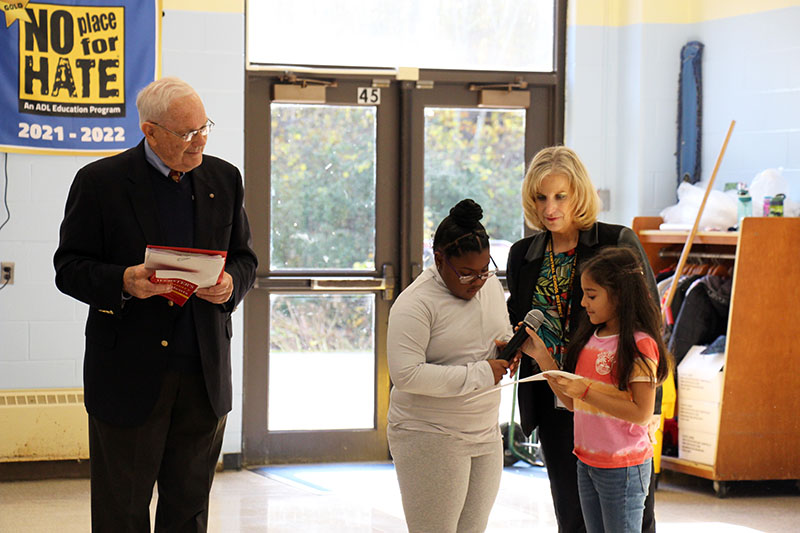 A man stands to the left while two third=grade girls read a thank you note to him, while a woman stands with them.