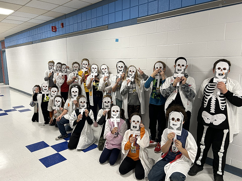 A large group of fourth grades students in two rows, front row squatting. They all have skeleton masks in front of their faces.