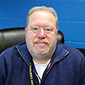 A man with short light hair wearing a blue zip sweatshirt sits in a black chair with a small smile.