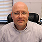 A man wearing glass and a button-down white shirt is sitting and looks in the camera.