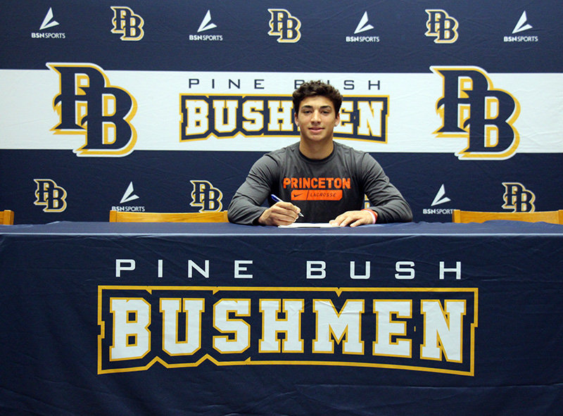 A young man sits at a table with a pen in hand to sign a document. He is smiling. The tablecloth says Pine Bush Bushmen.