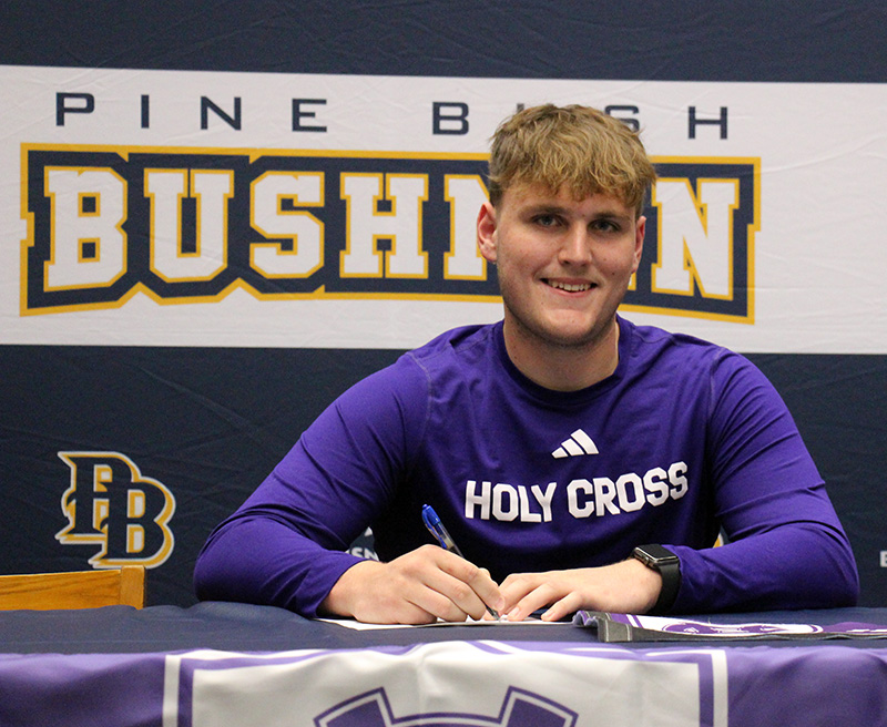 A young man smiles as he sits at a table. He is wearing a purple sweatshirt that says Holy Cross.