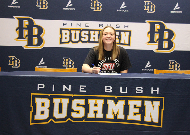 A young woman with long blonde hair sits at a tablethat says Pine Bush Bushmen. She is smiling.