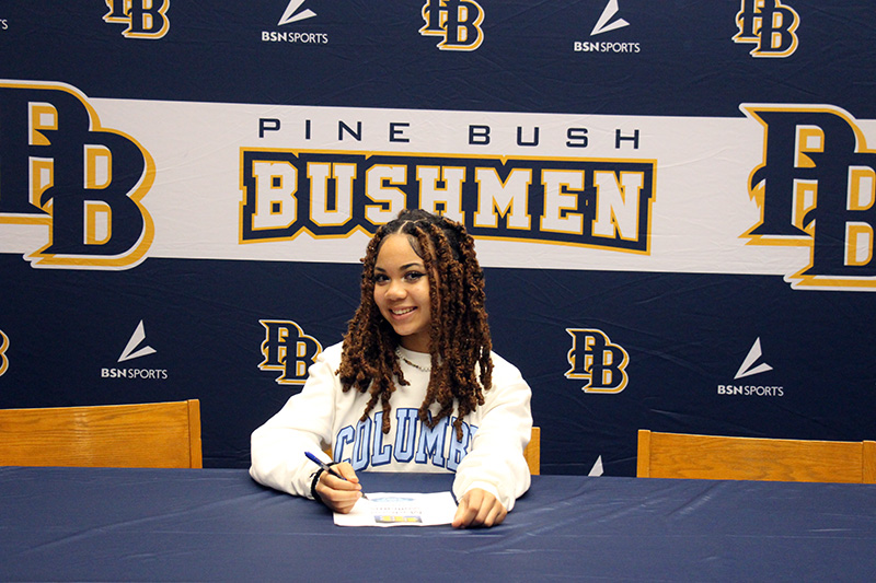 A young woman with long dark hair smiles as she sits with a pen in her hand. Behind her is a banner that says Pin eBush Bushmen in blue, gold and white.