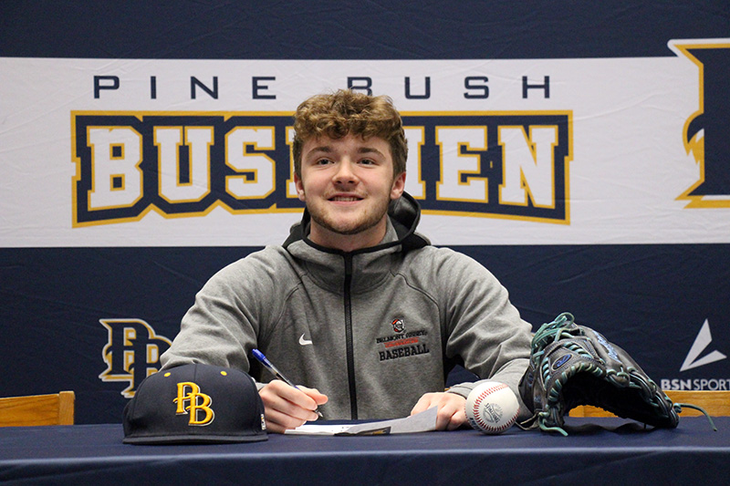 A young man smiles broadly as he sits at a table signing a paper. There are baseball hats and a baseball glove on the table.