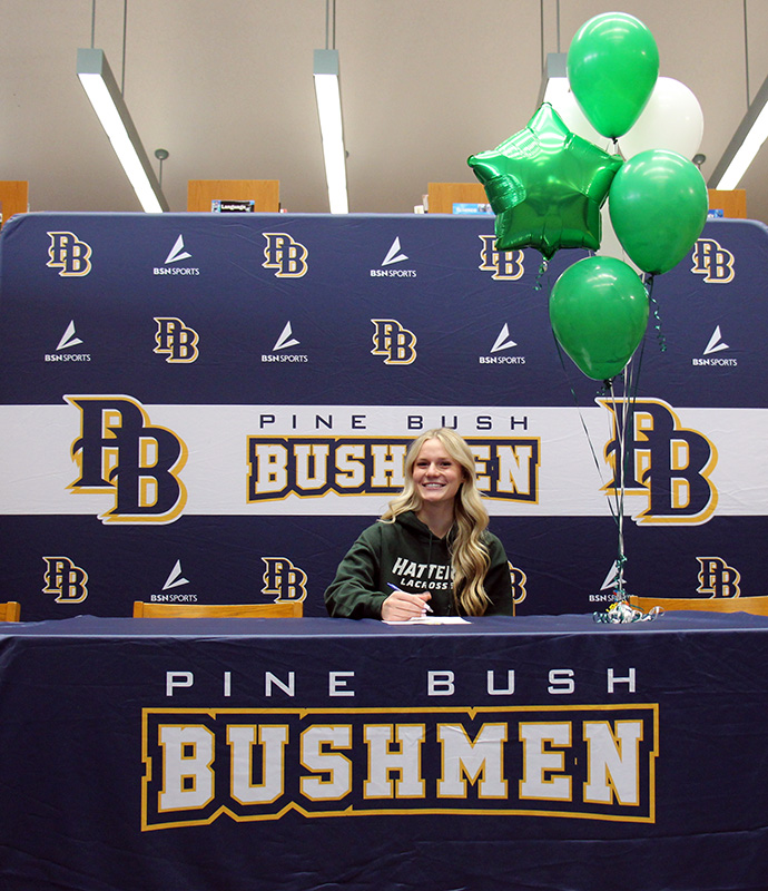 A young woman with long blonde hair smiles as she sits at a table. The tablecloth says Pin eBush bushmen and there are green balloons beside her.