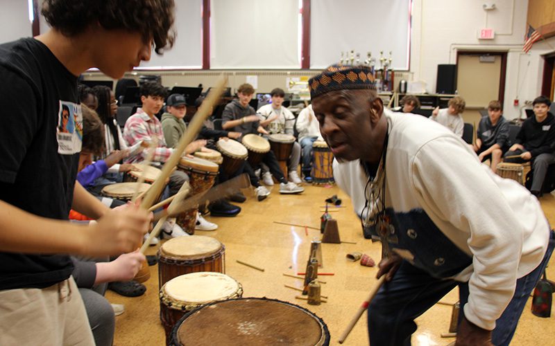 High school students line a room, all drumming with hands or sticks. A man in front of the room leans in to one of the drummers.
