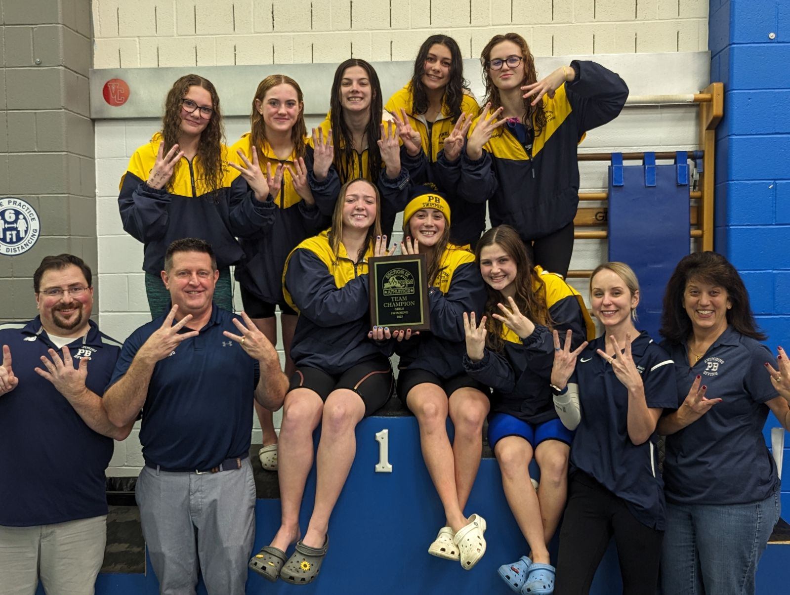 A group of eight high school swimmers, with wet hair, and four coaches all smiling. They are all holding up six fingers.