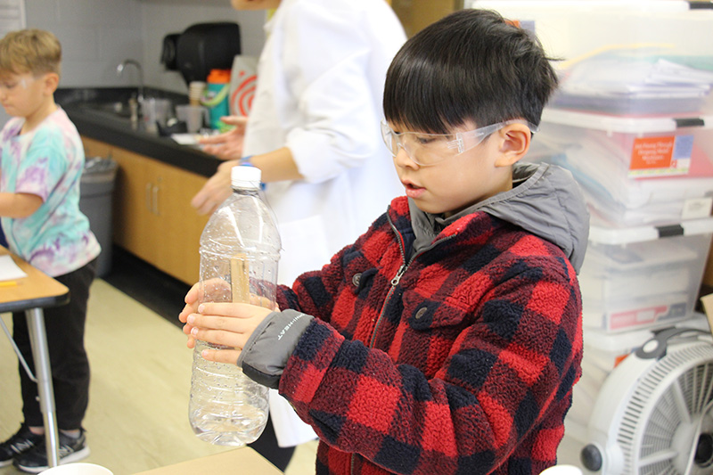 An elementary-age boy with short black hair wearing a black and white check shirt holds a clear plastic bottle and presses it.