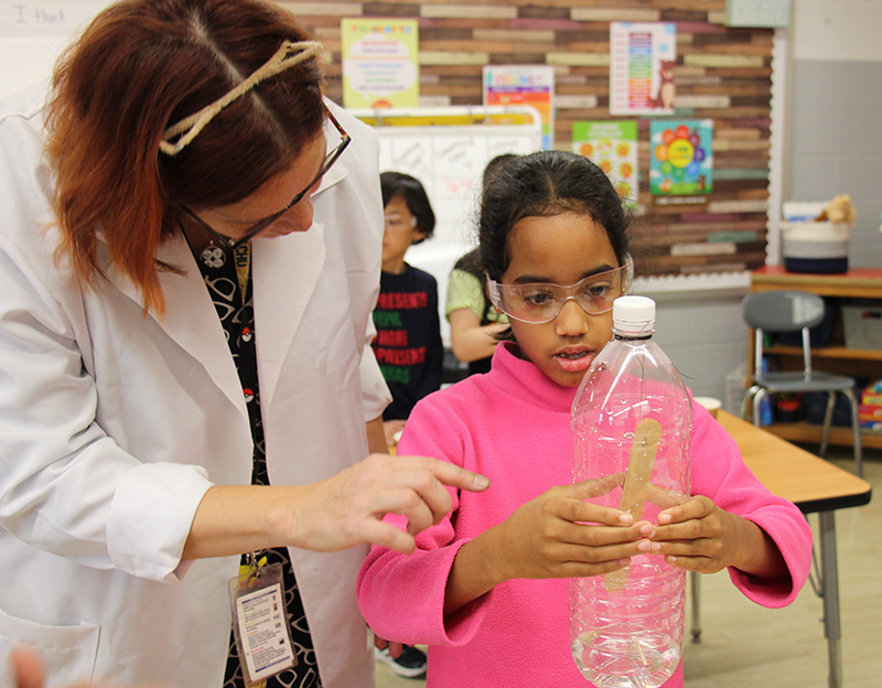 A girl in a pink sweater holds a clear plastic bottle and presses it while a woman wearing a white lab coat stands next to her.