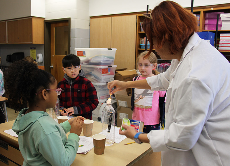 A woman in a white labcoat drops a lit match into a clear plastic bottle that has water in it. Three third-graders watch as she does it.