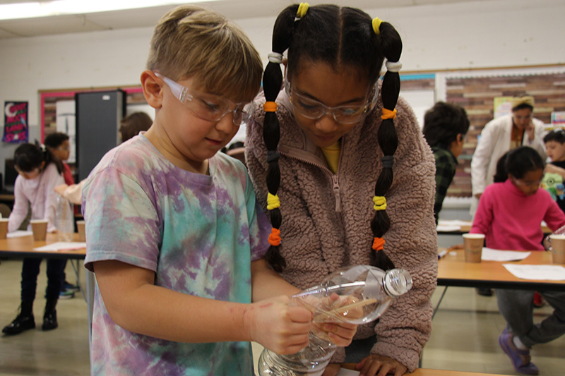 Two third-graders watch as one squeezes a clear plastic bottle doing an experiment.