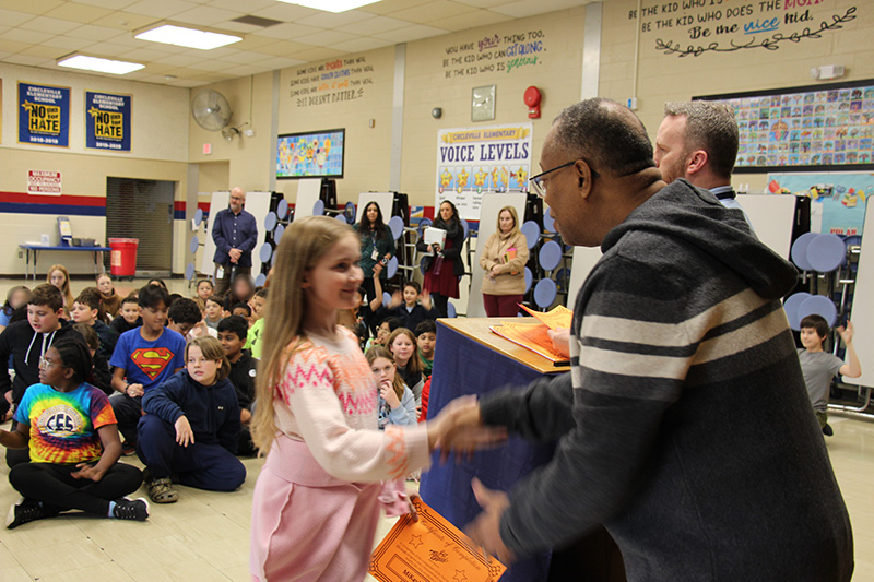 A fifth-grade girl shakes hands with a man. There are many kids behind them.