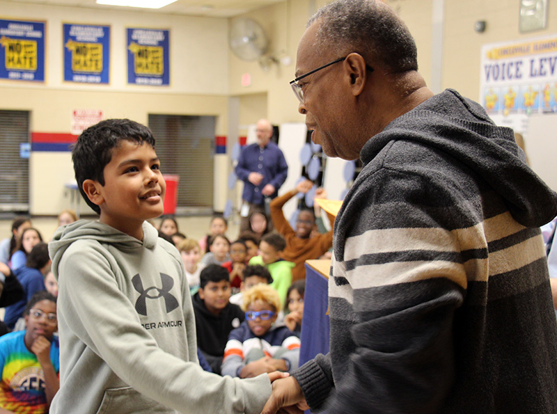A fifth-grade boy shakes hands with a man. There are many kids behind them.