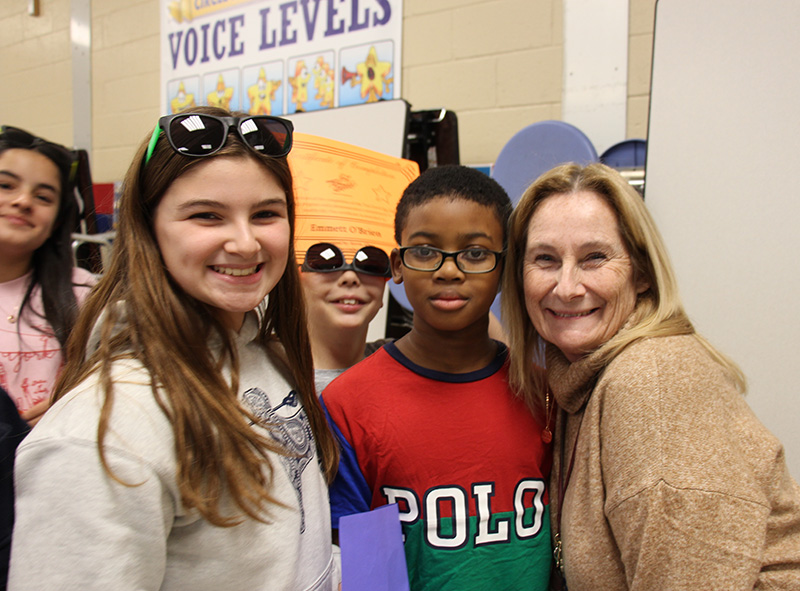 Three fifth-grade students smile as they stand with a woman with mid-length blonde hair. All are smiling.