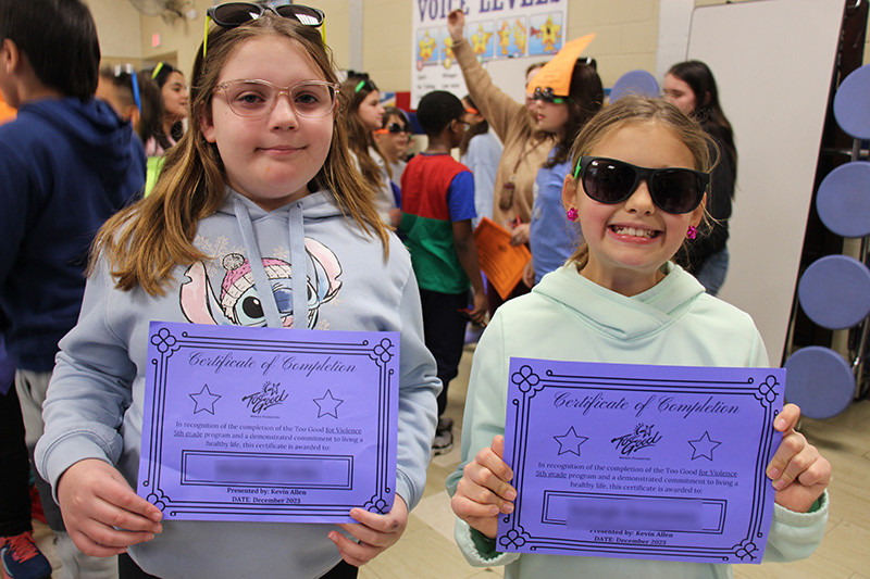 Two fifth-grade girls smile and hold purple certificates in front of them.