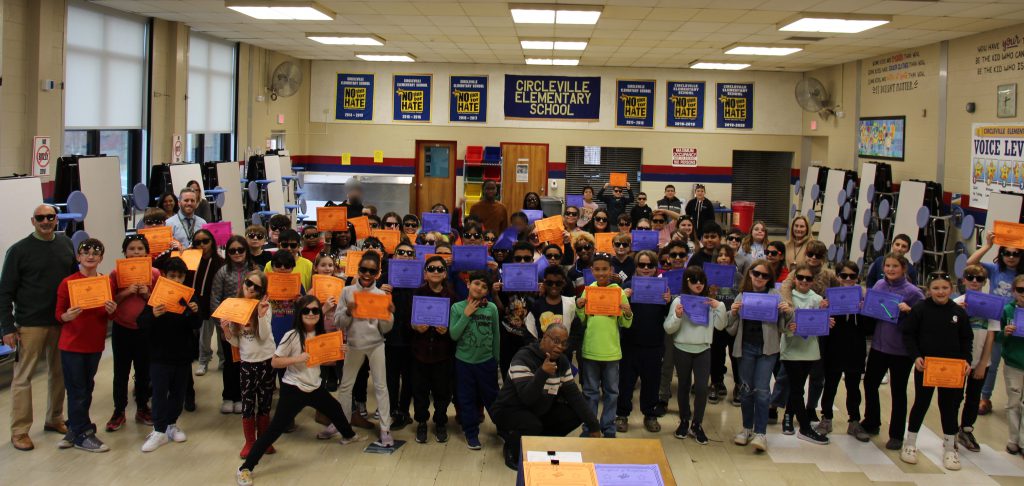 A group of about 80 fifth-grade students stand together in a cafeteria holding up their certificates. There are a couple of adults mixed in.