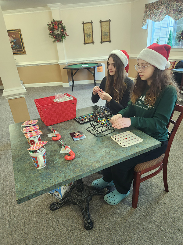 Two middle school girls wearing red and white santa hats sit at a table pulling numbers for a bingo game.