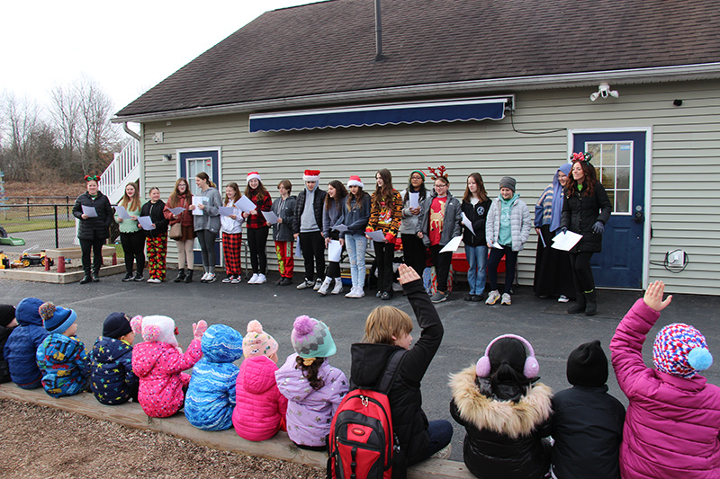A line of middle school students stand against a building and sing to little kids sitting in a row.