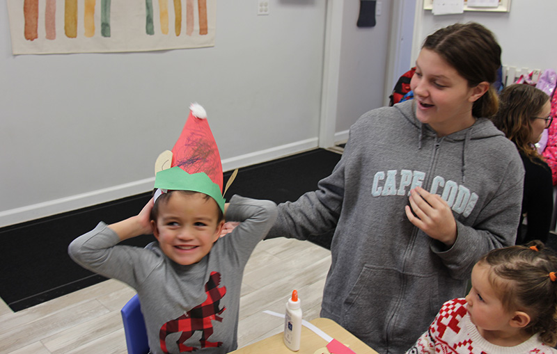 A little boy smiles wearing a red and green santa hat he made. There is a middle school kid next to him. 