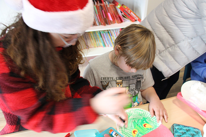 A middle school girl with long dark hair wearing a red and white santa hat helps a boy color a grinch picture.