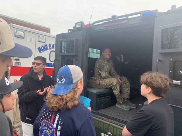 A group of eighth-grade kids stand looking into an armored military vehicle. There is a man in fatigues sitting inside of it.