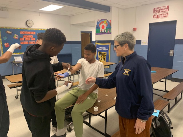 A kid sits on a table as another kid uses tape to wrap his arm. A woman with short hair, wearing a blue sweatshirt looks on and instructs them.