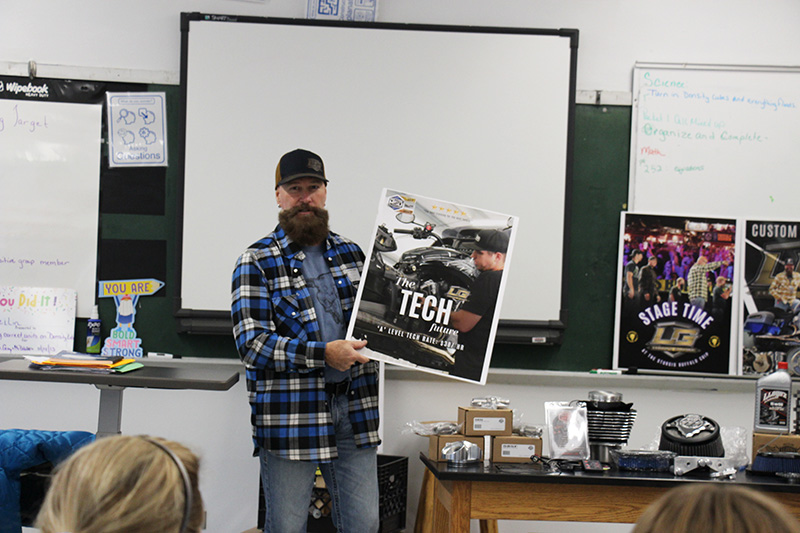 A man with a plaid shirt and hat, full beard, holds up a sign that says Tech on it with a picture of a motorcycle in the background.