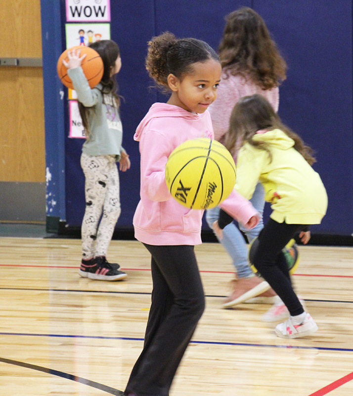 A second-grade girl with her dark hair pulled back, wearing a pink sweatshirt and black pants, dribbles a yellow basketball.
