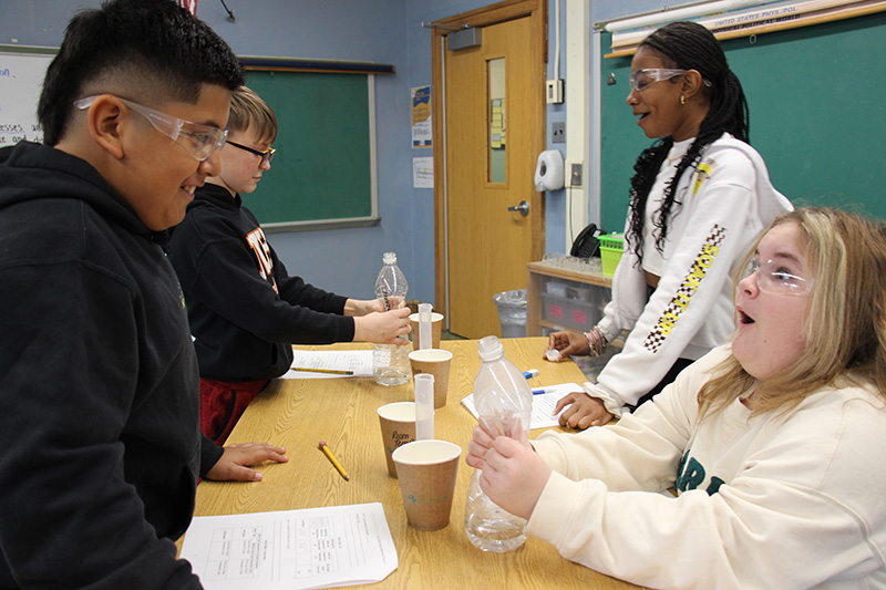 Third-grade students work together on a science project with a clear plastic bottle. They are squeezing the bottle. 