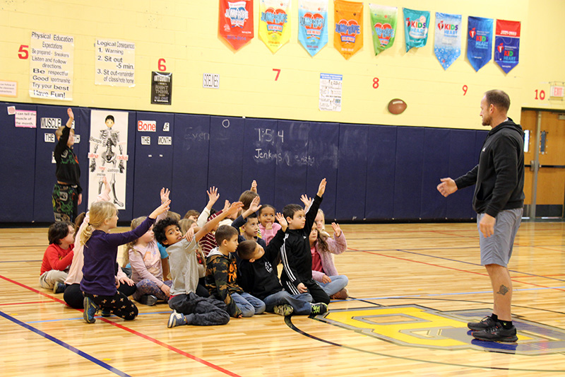A group of second-grade kids sit on a gym floor, many raising their hands as their teacher, a man in a sweatshirt and shorts, stands to the right.