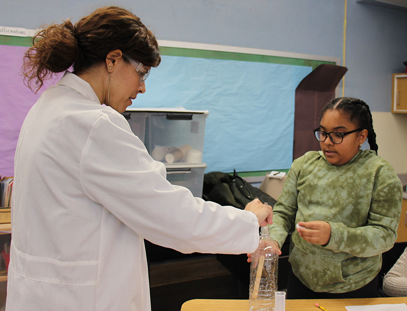 A woman in a white coat helps a third-grade student with her experiment.
