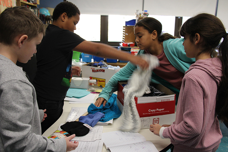 Fourth-grade students, two boys and two girls, lay winter hats, gloves, scarves and mittens  on a measuring stick.