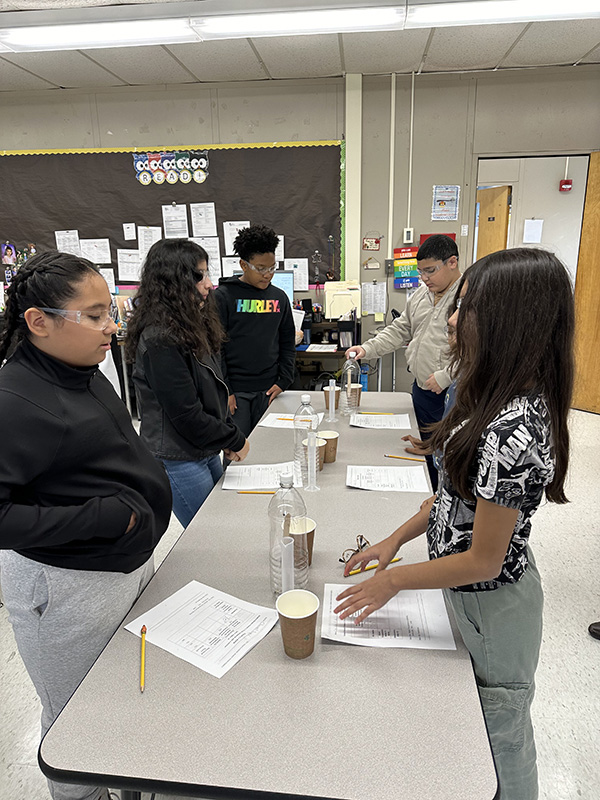 A group of six fifth-grade students work at a table on a science experiment that involves a tall bottle.