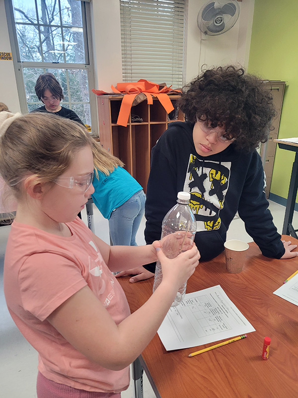 Two students work together on a science experiment. One is squeezing a clear plastic water bottle.
