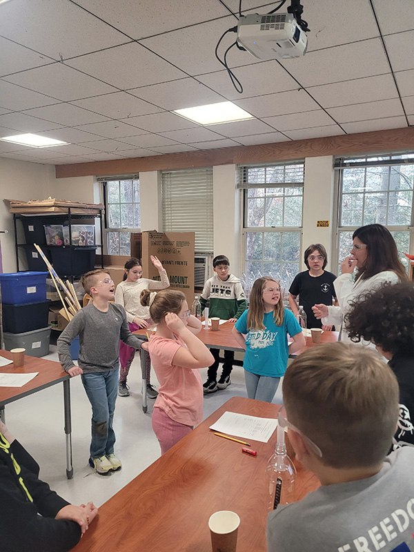 A class of students listen as a woman describes their science lesson to them.